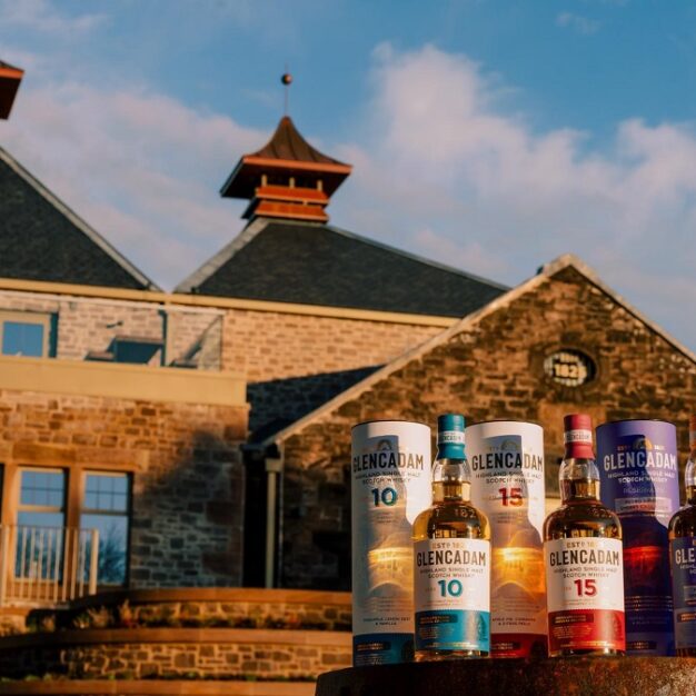 A close-up of three Glencadam Highland Single Malt Scotch Whisky bottles (10-year, 15-year, and Reserva de Jerez) and their decorative canisters, positioned in front of the distillery's stone buildings.