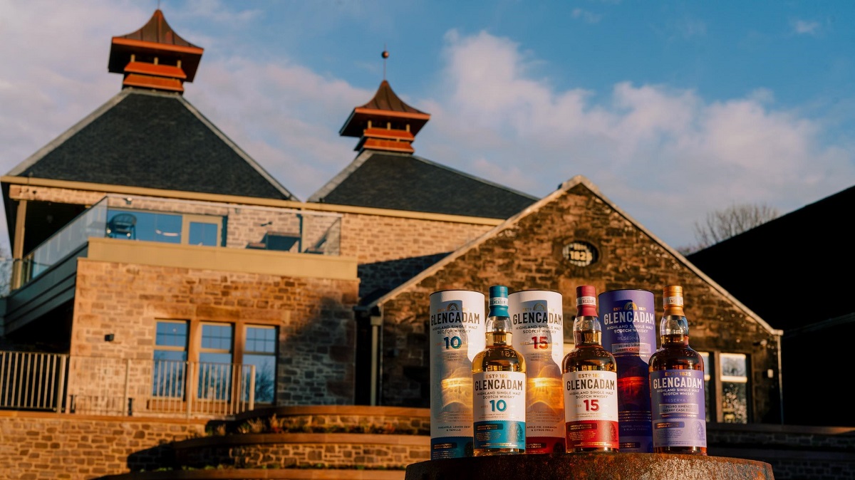 A close-up of three Glencadam Highland Single Malt Scotch Whisky bottles (10-year, 15-year, and Reserva de Jerez) and their decorative canisters, positioned in front of the distillery's stone buildings.