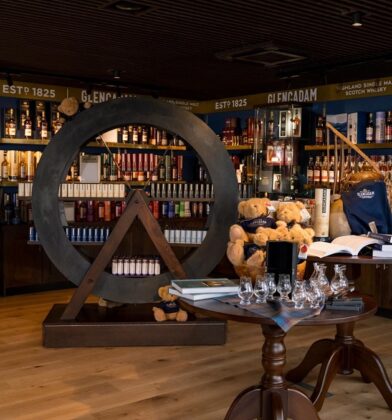 A well-lit gift shop area with wooden floors, featuring a large circular wooden shelf display, tables with glassware and books, and branded apparel on wall racks.