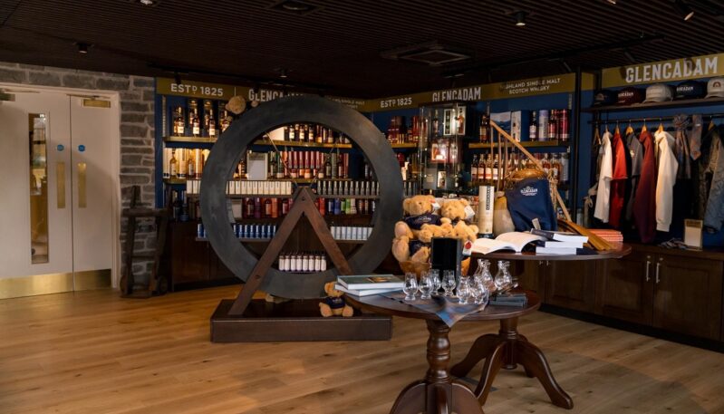 A well-lit gift shop area with wooden floors, featuring a large circular wooden shelf display, tables with glassware and books, and branded apparel on wall racks.