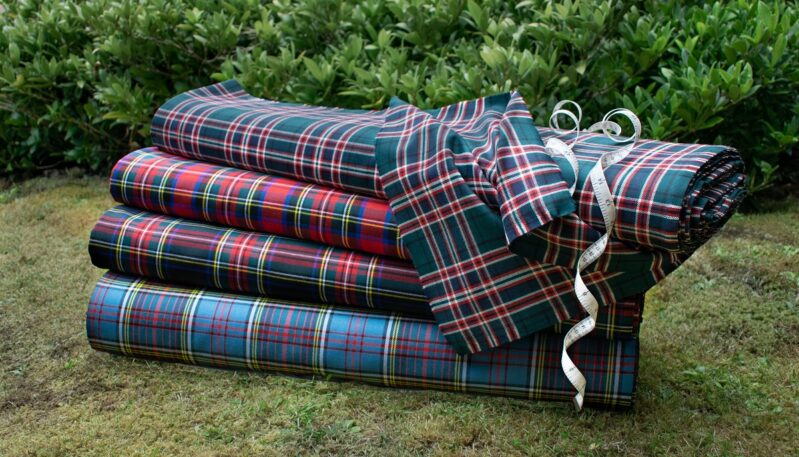 A stack of four tartan fabric rolls in various colors (green, red, dark red, and blue) lying on mossy grass, with a measuring tape draped over the top roll.