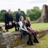 Four people posing on a stone wall in front of old stone ruins and trees; two men in suits and kilts stand behind two women seated and wearing tartan skirts.