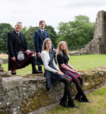 Four people posing on a stone wall in front of old stone ruins and trees; two men in suits and kilts stand behind two women seated and wearing tartan skirts.