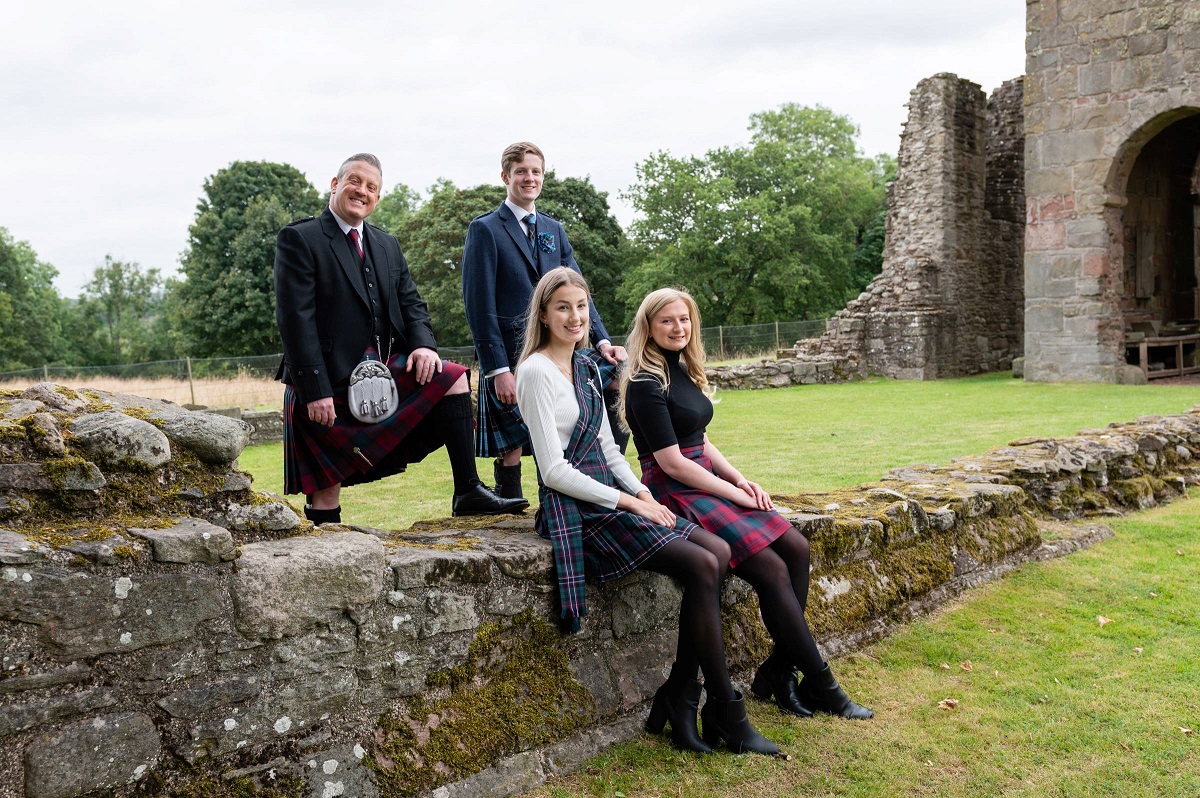 Four people posing on a stone wall in front of old stone ruins and trees; two men in suits and kilts stand behind two women seated and wearing tartan skirts.
