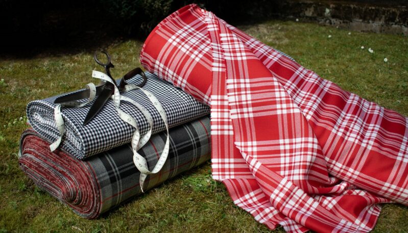 Three rolls of fabric on the grass—one in red and white tartan, one in black and white houndstooth, and one in a dark gray tartan—with a pair of vintage scissors and a measuring tape resting on the rolls.