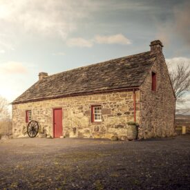 A traditional stone cottage with a slate roof, red-painted wooden door, and red window frames, featuring a wooden wagon wheel leaning against the exterior wall.