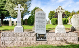 The gravesite of J.M. Barrie and his family, featuring stone gravestones and Celtic crosses enclosed within a low stone wall set against a sunny, green cemetery backdrop.