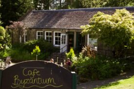 The entrance to Cafe Byzantium, featuring a handwritten sign in the foreground and a rustic stone building with outdoor seating and lush greenery in the background.
