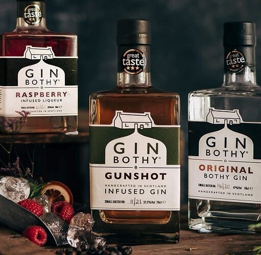 Three bottles of Gin Bothy products—Raspberry Infused Liqueur, Gunshot Infused Gin, and Original Bothy Gin—are arranged on a dark, rustic surface. The bottles feature "Great Taste" award stickers and are accompanied by fresh raspberries, ice in a metal scoop, and sprigs of heather against a dark, moody background.