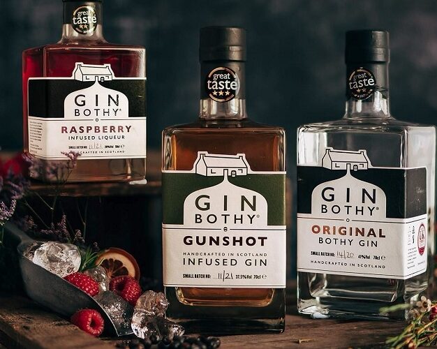 Three bottles of Gin Bothy products—Raspberry Infused Liqueur, Gunshot Infused Gin, and Original Bothy Gin—are arranged on a dark, rustic surface. The bottles feature "Great Taste" award stickers and are accompanied by fresh raspberries, ice in a metal scoop, and sprigs of heather against a dark, moody background.
