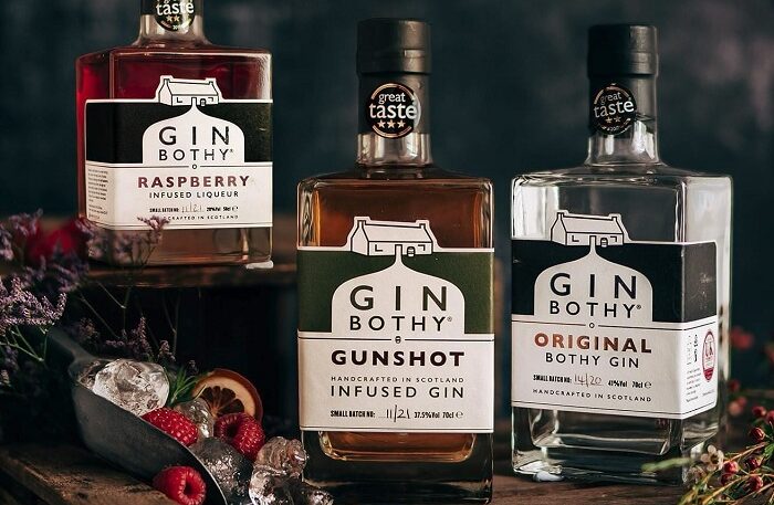 Three bottles of Gin Bothy products—Raspberry Infused Liqueur, Gunshot Infused Gin, and Original Bothy Gin—are arranged on a dark, rustic surface. The bottles feature "Great Taste" award stickers and are accompanied by fresh raspberries, ice in a metal scoop, and sprigs of heather against a dark, moody background.