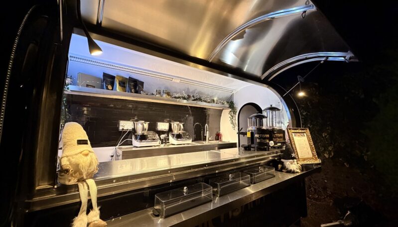 A sleek, black food trailer named "Bella" is illuminated at night, featuring a stainless steel service counter and a curved metal roof. The interior is equipped with professional hot chocolate dispensers and mixing machines, while festive gnomes and a gold-framed menu sit on the counter.