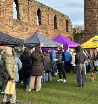 A bustling outdoor market scene with people gathered on a grassy area in front of the tall, weathered red-stone ruins of a historic building. Several vendor tents in black, gray, purple, and yellow are set up, with the yellow tent bearing the words "Eat • Buzz • Repeat." Shoppers, many wearing winter coats, browse the stalls. A small dog on a leash is visible on the right.