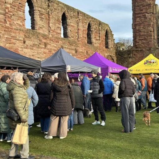 A bustling outdoor market scene with people gathered on a grassy area in front of the tall, weathered red-stone ruins of a historic building. Several vendor tents in black, gray, purple, and yellow are set up, with the yellow tent bearing the words "Eat • Buzz • Repeat." Shoppers, many wearing winter coats, browse the stalls. A small dog on a leash is visible on the right.