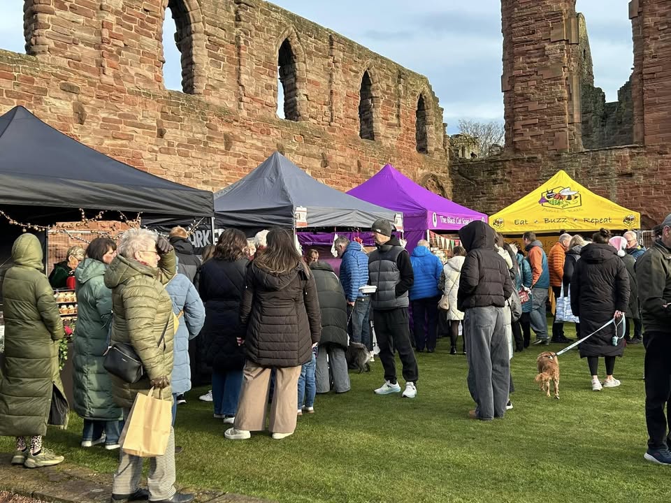 A bustling outdoor market scene with people gathered on a grassy area in front of the tall, weathered red-stone ruins of a historic building. Several vendor tents in black, gray, purple, and yellow are set up, with the yellow tent bearing the words "Eat • Buzz • Repeat." Shoppers, many wearing winter coats, browse the stalls. A small dog on a leash is visible on the right.
