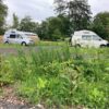 A slightly low-angle outdoor shot showing two campervans or motorhomes parked in a grassy, partially gravel area. In the foreground, there is a dense patch of weeds, wildflowers, and grass. Two high-top vans are visible in the background: one light gray/silver on the left and one white on the right. A wooden fence and trees form the boundary of the parking area, suggesting a dedicated stopover or aire.