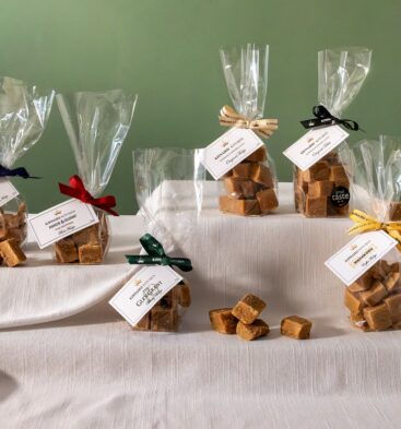 Six small clear bags filled with squares of fudge or tablet are arranged on a white cloth-covered display. Each bag is tied with a different colored ribbon—dark blue, red, dark green, cream, black, and yellow—and features a white label from "Kinnaird Kitchen" identifying the specific flavor, such as Spiced Rum and Raisin or Glen Grant Whisky. A few loose pieces of fudge sit in the center of the display against a muted green wall.