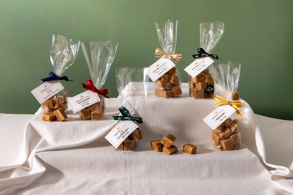 Six small clear bags filled with squares of fudge or tablet are arranged on a white cloth-covered display. Each bag is tied with a different colored ribbon—dark blue, red, dark green, cream, black, and yellow—and features a white label from "Kinnaird Kitchen" identifying the specific flavor, such as Spiced Rum and Raisin or Glen Grant Whisky. A few loose pieces of fudge sit in the center of the display against a muted green wall.