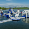 Large inflatable aqua park floating on a loch, featuring climbing walls, slides, balance obstacles and tunnels, with participants in buoyancy aids and rolling green countryside in the background.