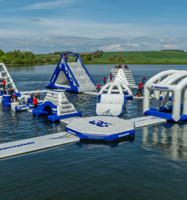 Large inflatable aqua park floating on a loch, featuring climbing walls, slides, balance obstacles and tunnels, with participants in buoyancy aids and rolling green countryside in the background.