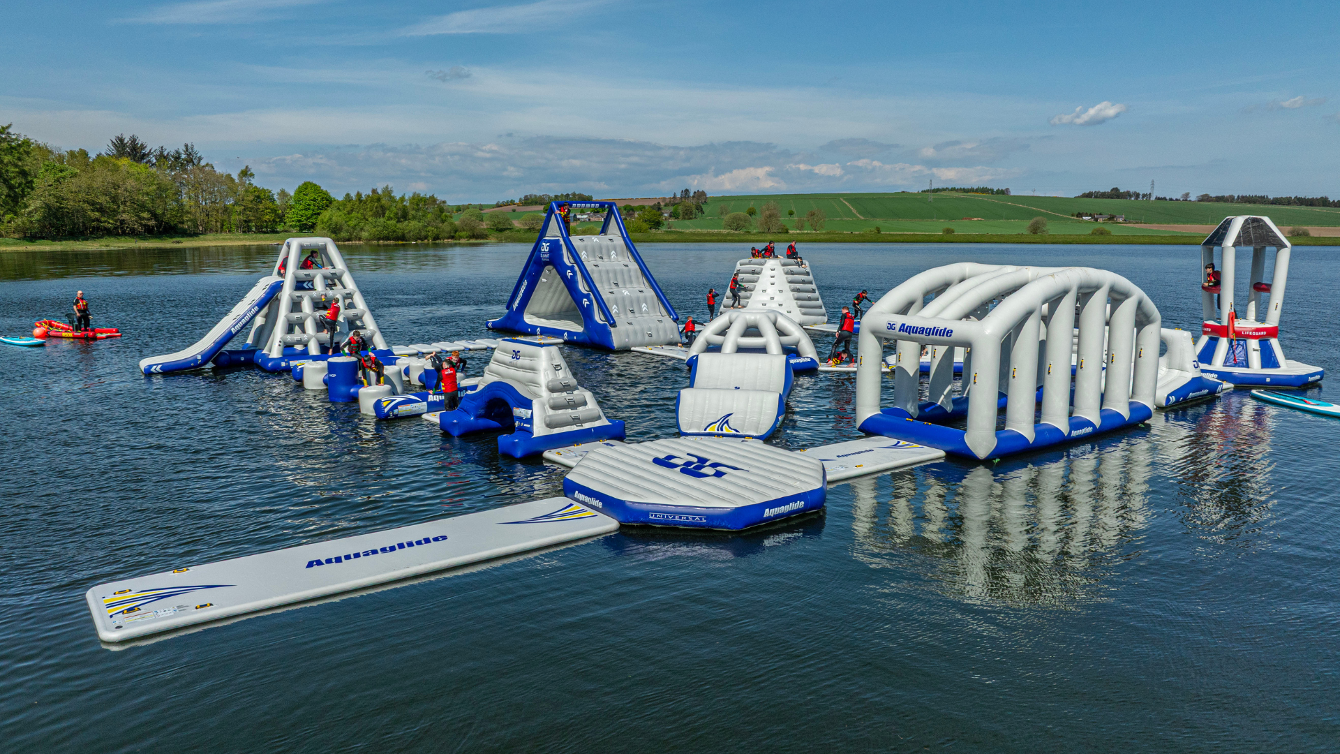 Large inflatable aqua park floating on a loch, featuring climbing walls, slides, balance obstacles and tunnels, with participants in buoyancy aids and rolling green countryside in the background.