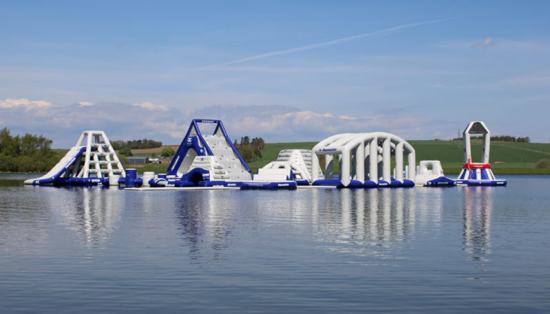 Large inflatable aqua park floating on a calm loch, featuring climbing towers, slides, tunnels and obstacle sections, with open countryside and a wide blue sky in the background.