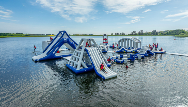 Visitors wearing buoyancy aids enjoying a floating inflatable obstacle course on a loch, with slides, climbing structures and balance challenges spread across the water against a rural landscape.