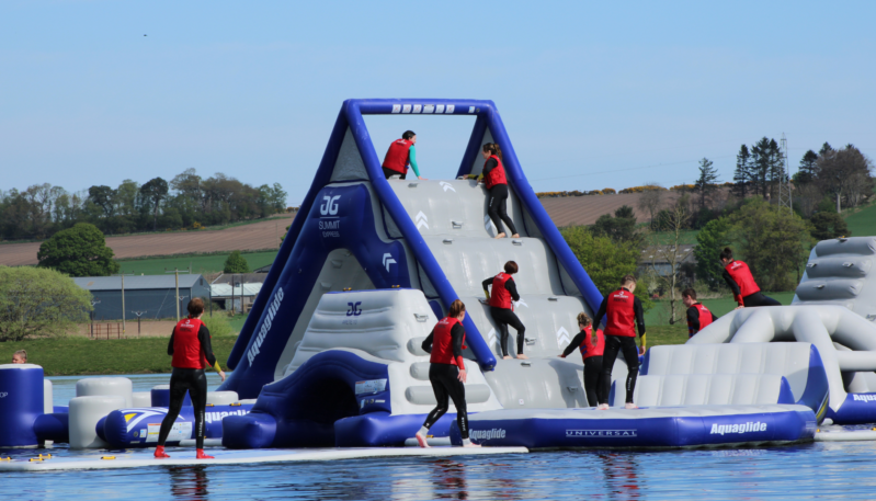 Visitors wearing red buoyancy aids climbing and crossing a giant inflatable obstacle course on a loch, centred around a tall blue-and-grey climbing tower with rural scenery behind.