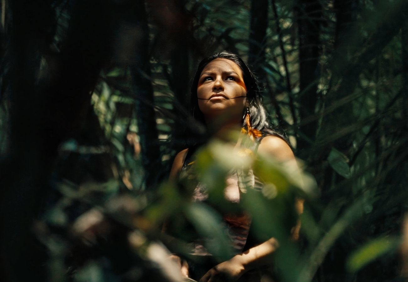 A person with long dark hair and traditional facial markings looks upward with a focused expression while partially obscured by dense forest foliage. They are adorned with orange feathered earrings, and warm sunlight filters through the canopy, creating dappled shadows across their face and the surrounding greenery.