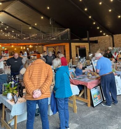 This image shows an indoor craft fair or market taking place in a rustic, barn-like venue with brick walls and a high black ceiling draped in warm string lights. Several people are browsing tables filled with handmade goods, including jewelry, framed artwork, and festive decorations. In the foreground, shoppers in casual winter jackets are viewed from behind as they inspect items, while vendors engage with customers across the display tables.