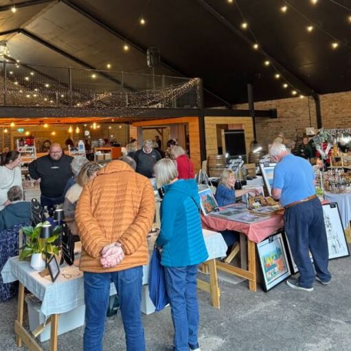 This image shows an indoor craft fair or market taking place in a rustic, barn-like venue with brick walls and a high black ceiling draped in warm string lights. Several people are browsing tables filled with handmade goods, including jewelry, framed artwork, and festive decorations. In the foreground, shoppers in casual winter jackets are viewed from behind as they inspect items, while vendors engage with customers across the display tables.