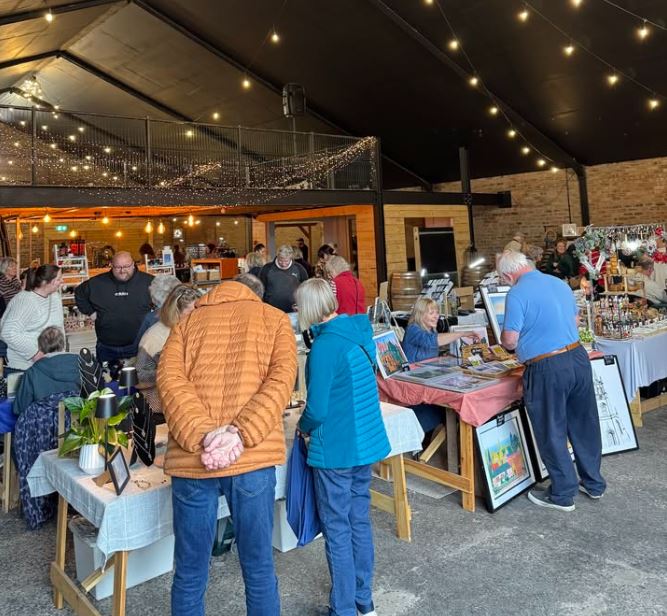 This image shows an indoor craft fair or market taking place in a rustic, barn-like venue with brick walls and a high black ceiling draped in warm string lights. Several people are browsing tables filled with handmade goods, including jewelry, framed artwork, and festive decorations. In the foreground, shoppers in casual winter jackets are viewed from behind as they inspect items, while vendors engage with customers across the display tables.