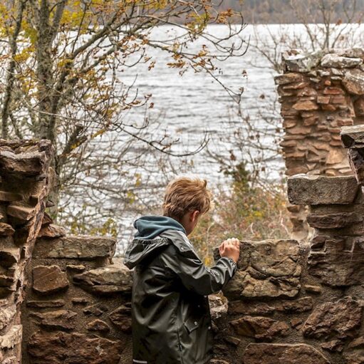 A child in a green jacket and blue hoodie is looking out from behind a stone wall. The stones are part of a ruined structure. Beyond the wall, a large body of water is visible, with trees on the far shore.