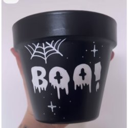 A hand holds a small black terra cotta pot decorated with white Halloween-themed illustrations. A spiderweb is on the upper left, and the word "BOO!" is painted in a drippy font across the front. White stars and sparkles are scattered around the pot.