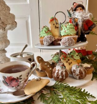 A festive photo of an afternoon tea set. In the foreground, a floral teacup on a saucer holds a cup of tea. Next to it, a two-tiered stand is filled with Christmas-themed treats, including cupcakes with green frosting, gingerbread men, and baked goods covered in white frosting. A small Santa Claus figurine stands at the top of the stand. A sprig of green leaves is arranged around the base of the stand.