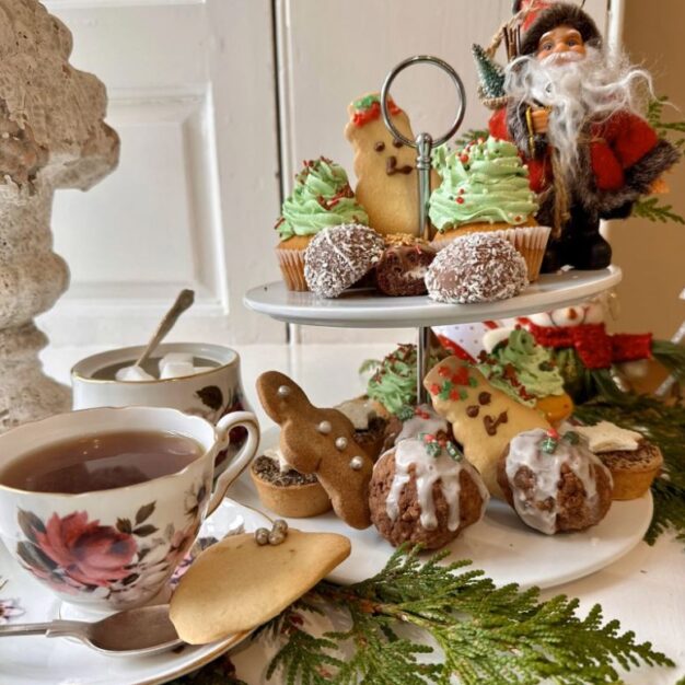 A festive photo of an afternoon tea set. In the foreground, a floral teacup on a saucer holds a cup of tea. Next to it, a two-tiered stand is filled with Christmas-themed treats, including cupcakes with green frosting, gingerbread men, and baked goods covered in white frosting. A small Santa Claus figurine stands at the top of the stand. A sprig of green leaves is arranged around the base of the stand.