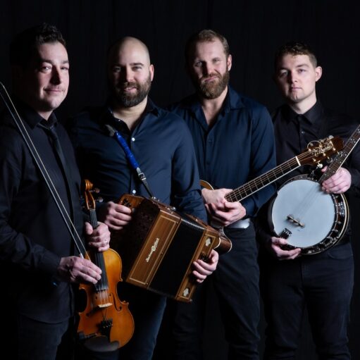 A professional portrait photograph of four men standing in a line against a black background. They are all wearing dark shirts and holding musical instruments: a violin and bow, a diatonic button accordion, a guitar, and a banjo.