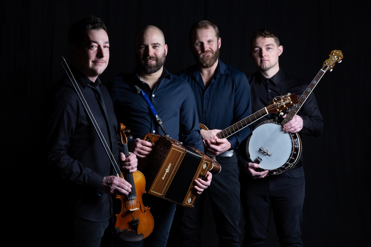 A professional portrait photograph of four men standing in a line against a black background. They are all wearing dark shirts and holding musical instruments: a violin and bow, a diatonic button accordion, a guitar, and a banjo.