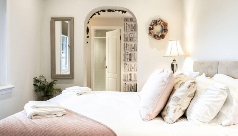 A bedroom with a large bed covered in white and pink bedding. A mirror hangs on the left wall, and a floral wreath is on the wall on the right. An arched doorway leads into another room with a door and a unique wallpaper that resembles bookshelves.