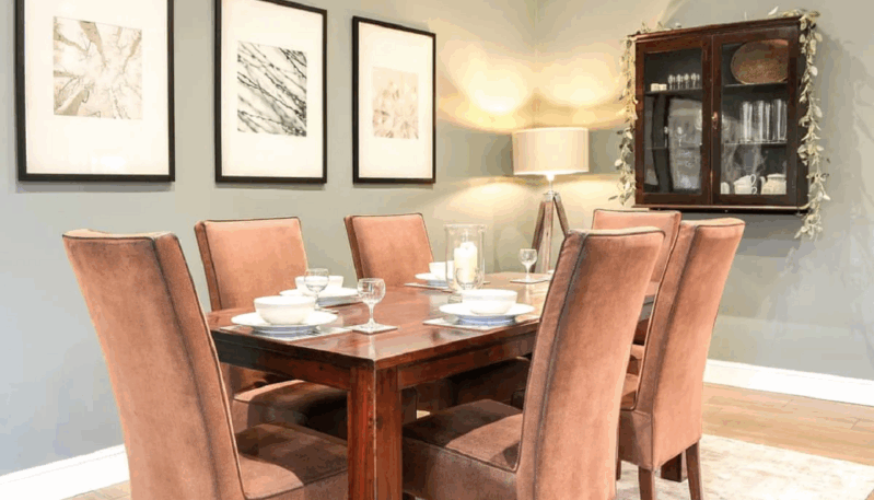 A dining area with a dark wood table set for six people. The table is surrounded by six upholstered chairs. The walls are a light sage green, and three framed pieces of art hang above a dark wood display cabinet on the wall. A lamp and a candle are on the table.
