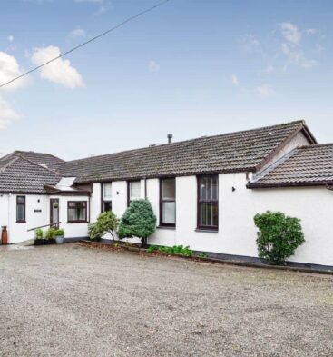 An exterior shot of a long, single-story white building with a tiled, reddish-brown roof. The building has multiple windows and a gravel driveway in front. A low hedge is on the left side of the building.