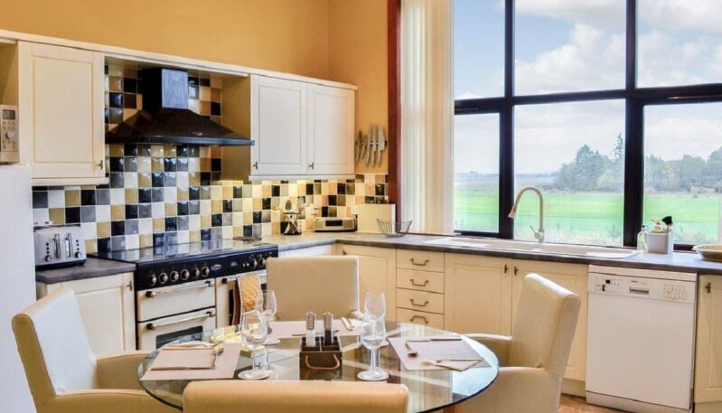 A bright kitchen with a large window that looks out onto a green field. The kitchen has off-white cabinets, a black stove, and a black and white tiled backsplash. A round glass table with four white upholstered chairs is in the center of the room.