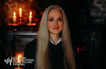 A medium-close portrait of a woman with long, straight blonde hair, a light complexion, and a slight smile. She is wearing a black jumper over a white collared shirt. In the dark background, three lit candles on a fireplace mantel are visible to the left. The logo for the Scottish International Storytelling Festival is in the bottom left corner.