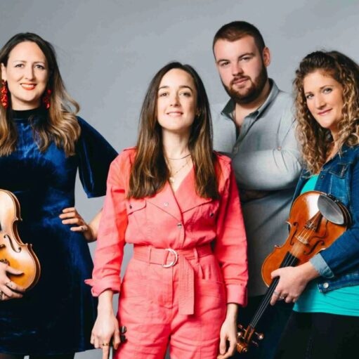 A studio portrait of four musicians standing in front of a gray background. From left to right: a woman in a dark blue dress holding a violin; a woman in a pink jumpsuit; a man in a gray shirt with his arms crossed; and a woman in a denim jacket holding a violin. They are all looking at the camera with confident, relaxed expressions.