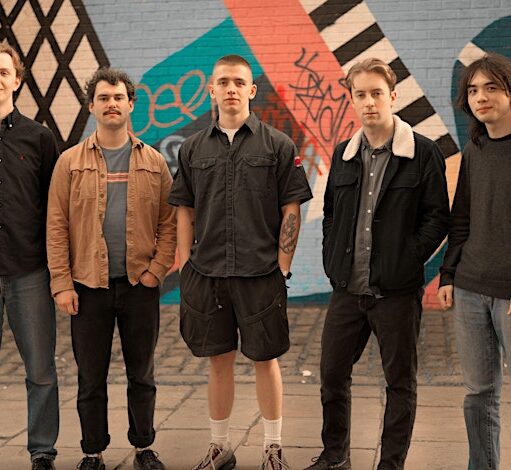 A full-body group portrait of five young men standing in a line in front of a brick wall and a colorful mural. From left to right: a man with a slight smile wearing a black button-down shirt and jeans; a man with a mustache and curly hair wearing a tan jacket over a t-shirt and dark pants with his hands in his pockets; a man in the center wearing a dark gray short-sleeved button-down shirt and shorts; a man wearing a black denim jacket with a white fleece collar and jeans; and a man with long, dark hair wearing a gray sweater and jeans. They all look straight at the camera.