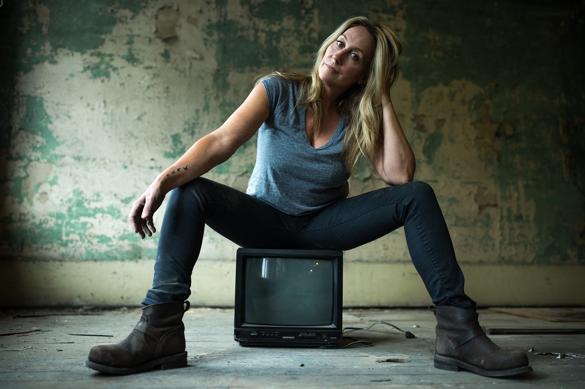 An image of singer-songwriter Amy Speace sitting on top of an old black television set. She is wearing a light blue-grey v-neck t-shirt and dark jeans. The background is a worn-out looking room with peeling paint on the walls.