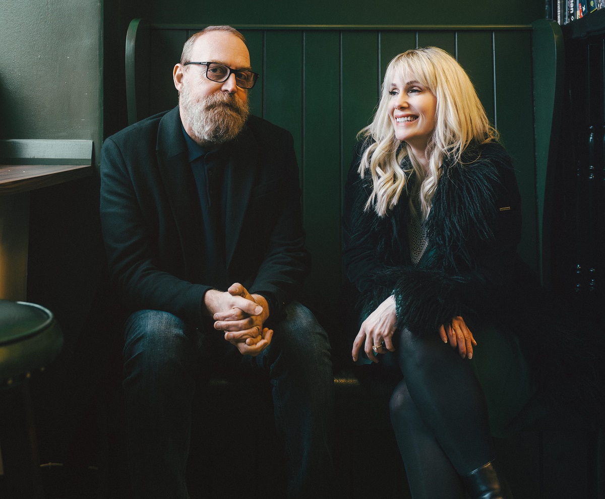 An image of two people, Heidi Talbot and Boo Hewerdine, sitting on a dark green bench indoors. Boo Hewerdine, on the left, has a beard and is wearing a black blazer and glasses. Heidi Talbot, on the right, has blonde hair and is wearing a black faux-fur jacket. Both are smiling.
