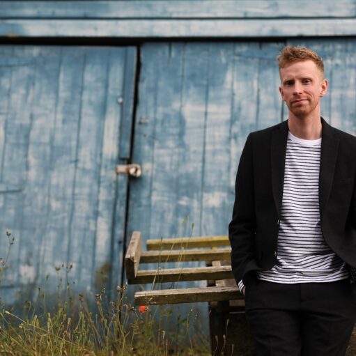 An image of singer-songwriter Adam Ross standing outdoors. He is wearing a black blazer over a striped t-shirt and black trousers. He is standing in front of two large, blue barn doors.