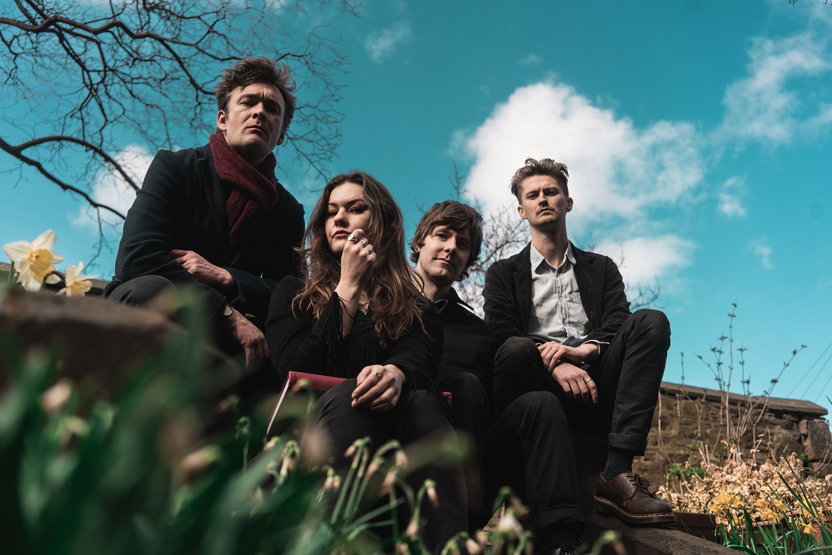An image of the band Kirsten Adamson and the Tanagers sitting outdoors. There are four people in the image. Two men are on the left side, and one man and one woman are on the right side. The person on the far left and the two people on the right are looking into the camera. The person in the front on the left is looking away from the camera. The sky is bright blue and there are yellow daffodils in the foreground.