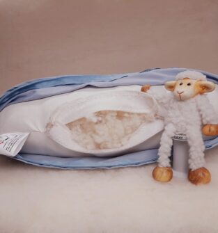 A rectangular pillow is shown with a light blue zippered cover and a white inner casing. The inner casing has a small slit revealing the fluffy wool filling. To the right of the pillow, a small toy sheep stands upright. The entire setup rests on a soft, woolly surface. A small label with a red sheep logo is visible on the side of the pillow.